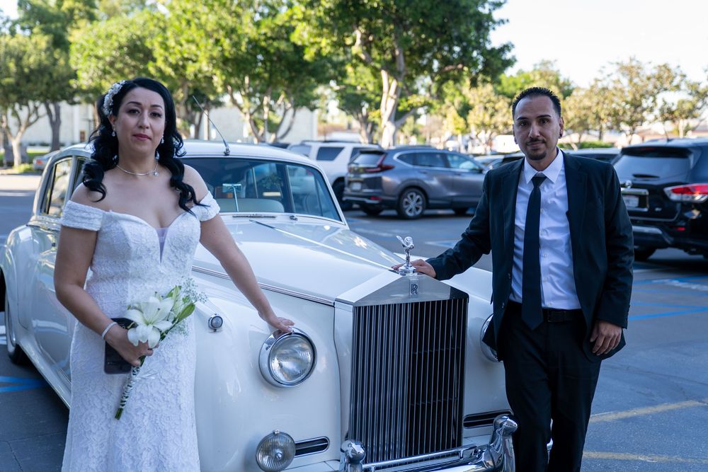 Bride and groom posing beside a classic white Rolls-Royce in a sunny parking lot on their wedding day.