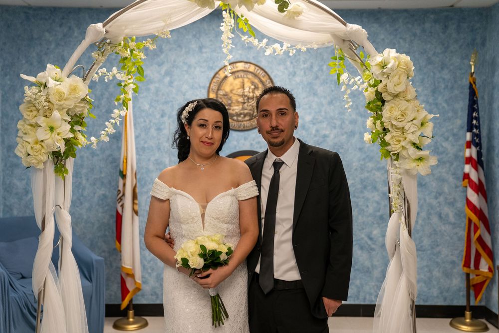 Couple in wedding attire posing under floral arch at courthouse ceremony with American flag.