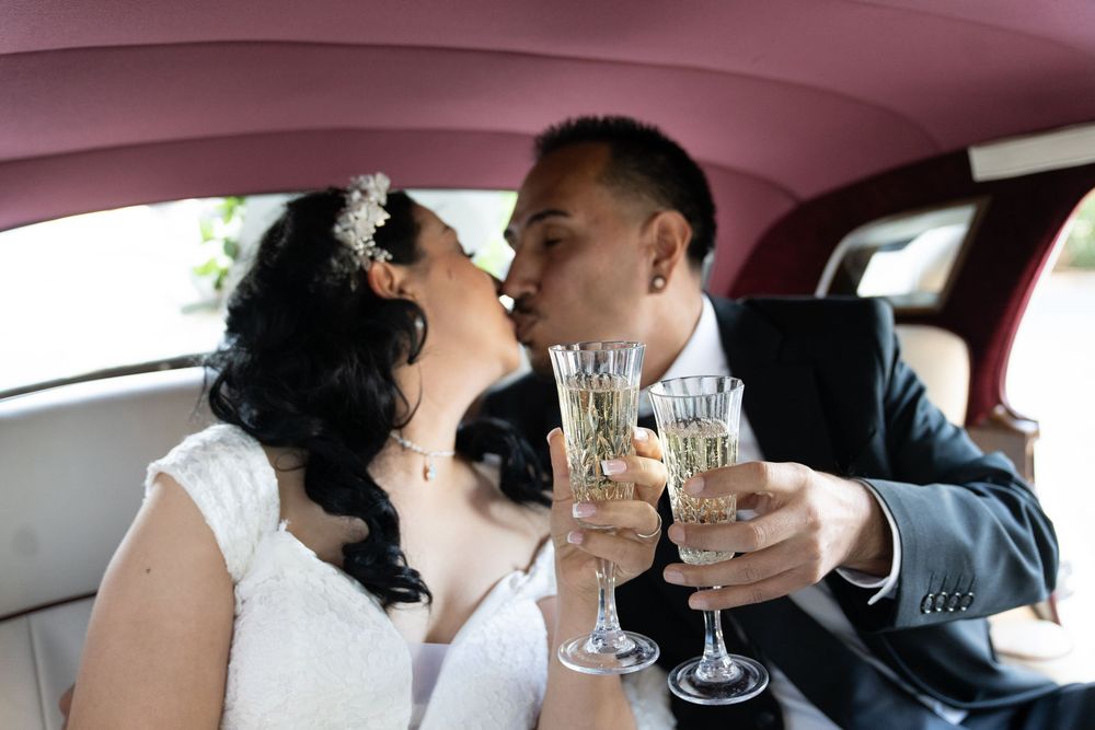 Newlywed couple kissing and toasting with champagne flutes in a pink limousine on their wedding day.