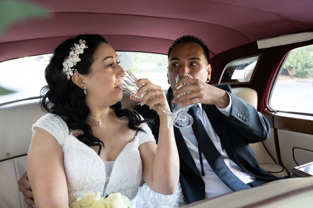 Bride and groom in wedding attire drinking champagne together in a luxury car on their wedding day.