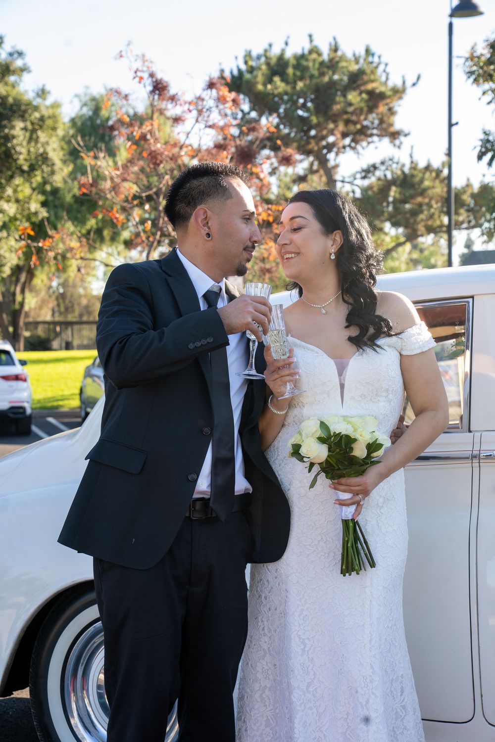 Bride and groom toasting with champagne glasses beside a vintage white car, bride holding a bouquet of white roses.