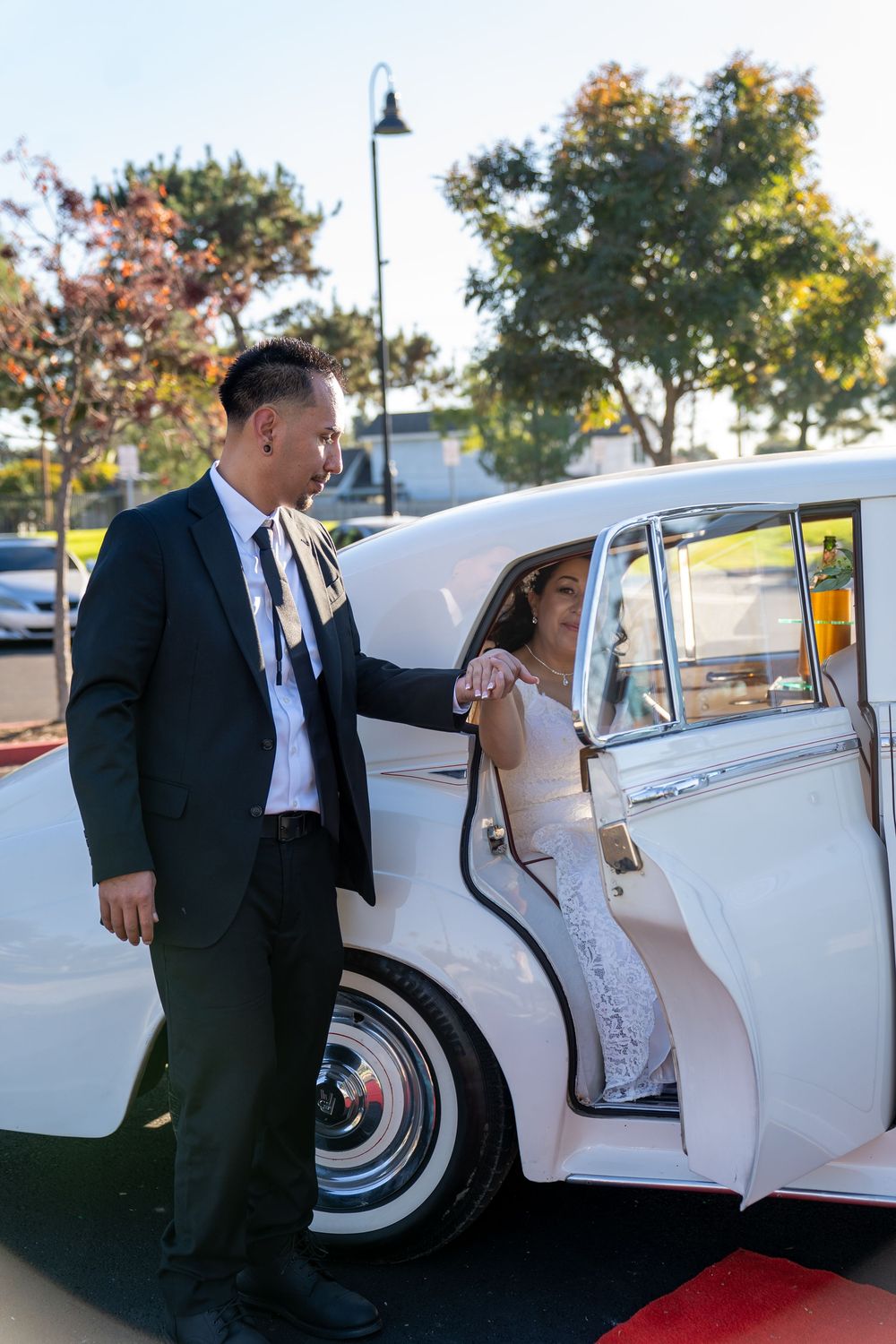 Groom in black suit helps bride in white lace dress exit a classic vintage white car on a red carpet.