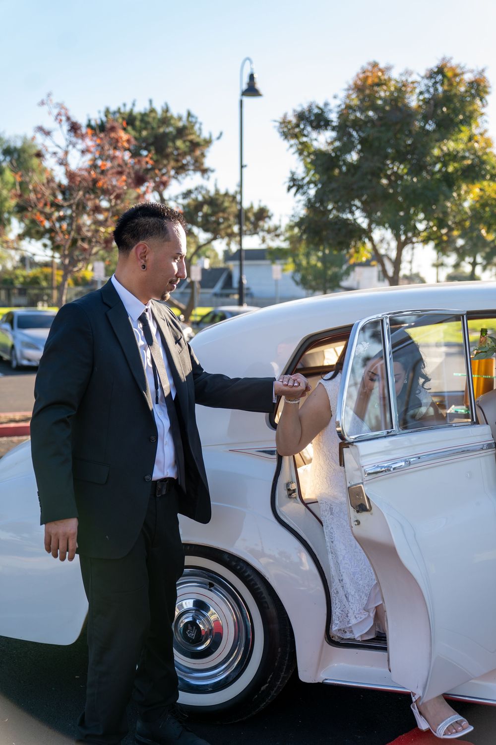 Groom in black suit opens vintage white car door for bride in white lace dress on wedding day.