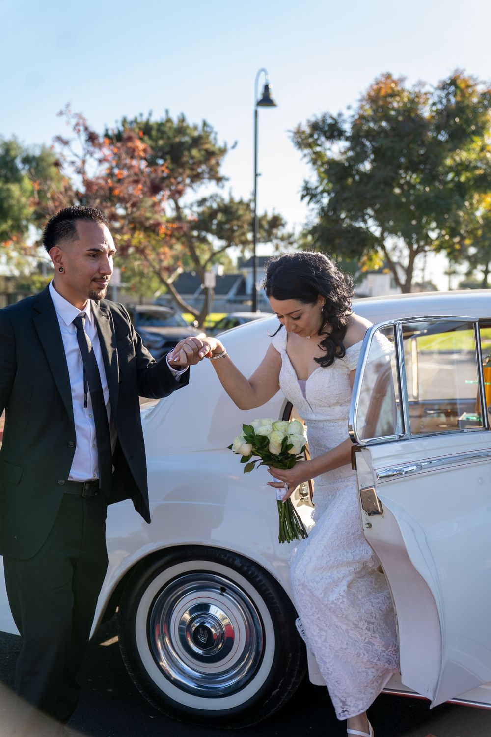Groom helps bride exit a vintage white car, bride holding white bouquet on a sunny wedding day.