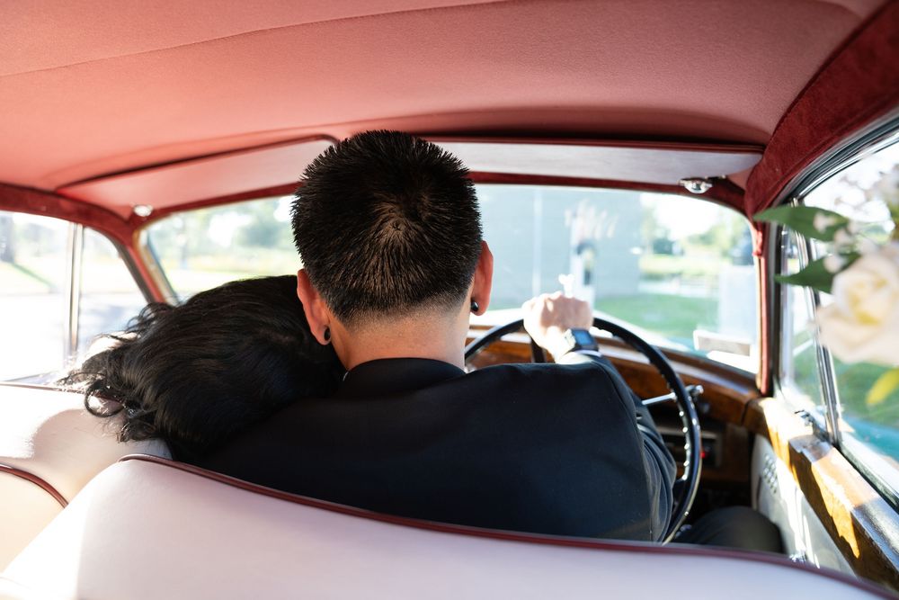 Couple embracing in a vintage classic car, viewed from behind, with a pink interior and retro steering wheel.