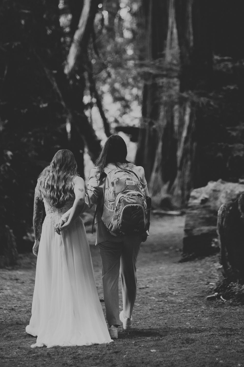 Black and white photo of two women walking through a forest, one in a wedding dress and one with a backpack.