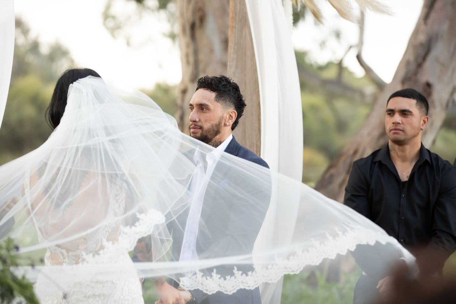 Groom in navy suit watches bride's veil flow during outdoor wedding ceremony with guest seated behind.