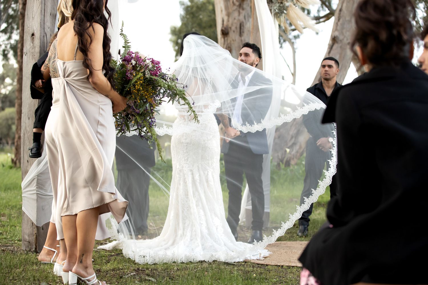 Bride in lace gown with flowing veil outdoors at garden wedding ceremony surrounded by guests.