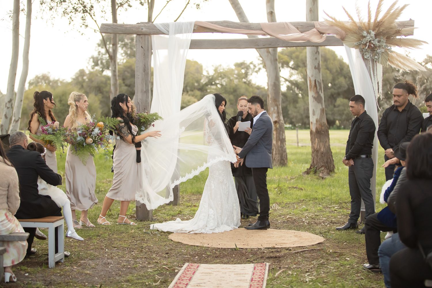 Outdoor wedding ceremony under rustic wooden arch with flowing white fabric, bride and groom exchanging vows on grass.