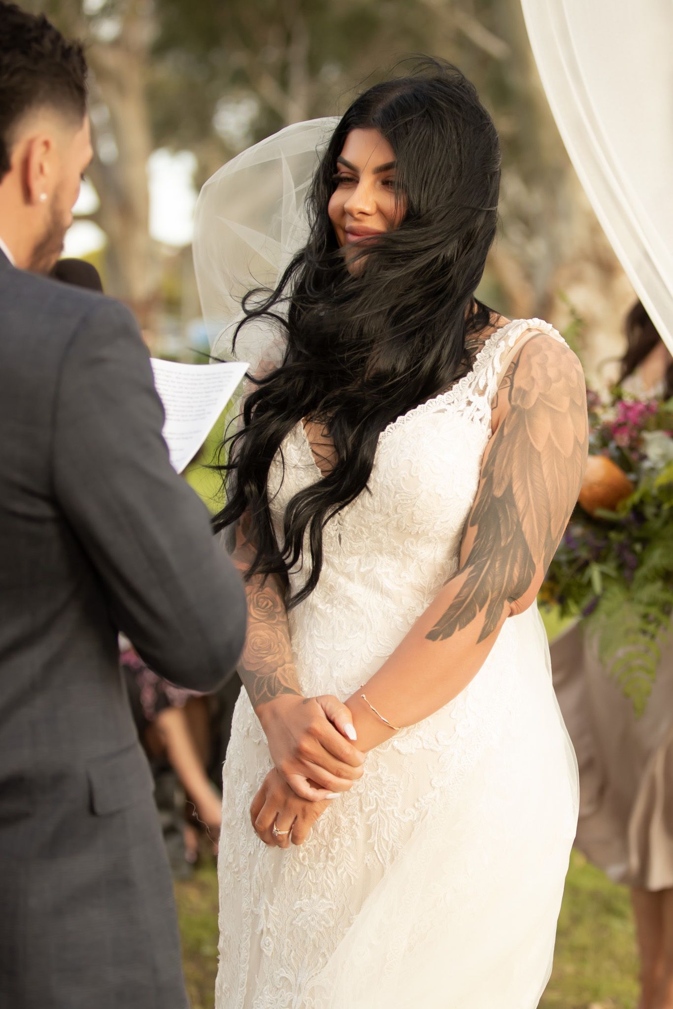 Tattooed bride in lace wedding gown smiles during outdoor ceremony while groom holds her hands.