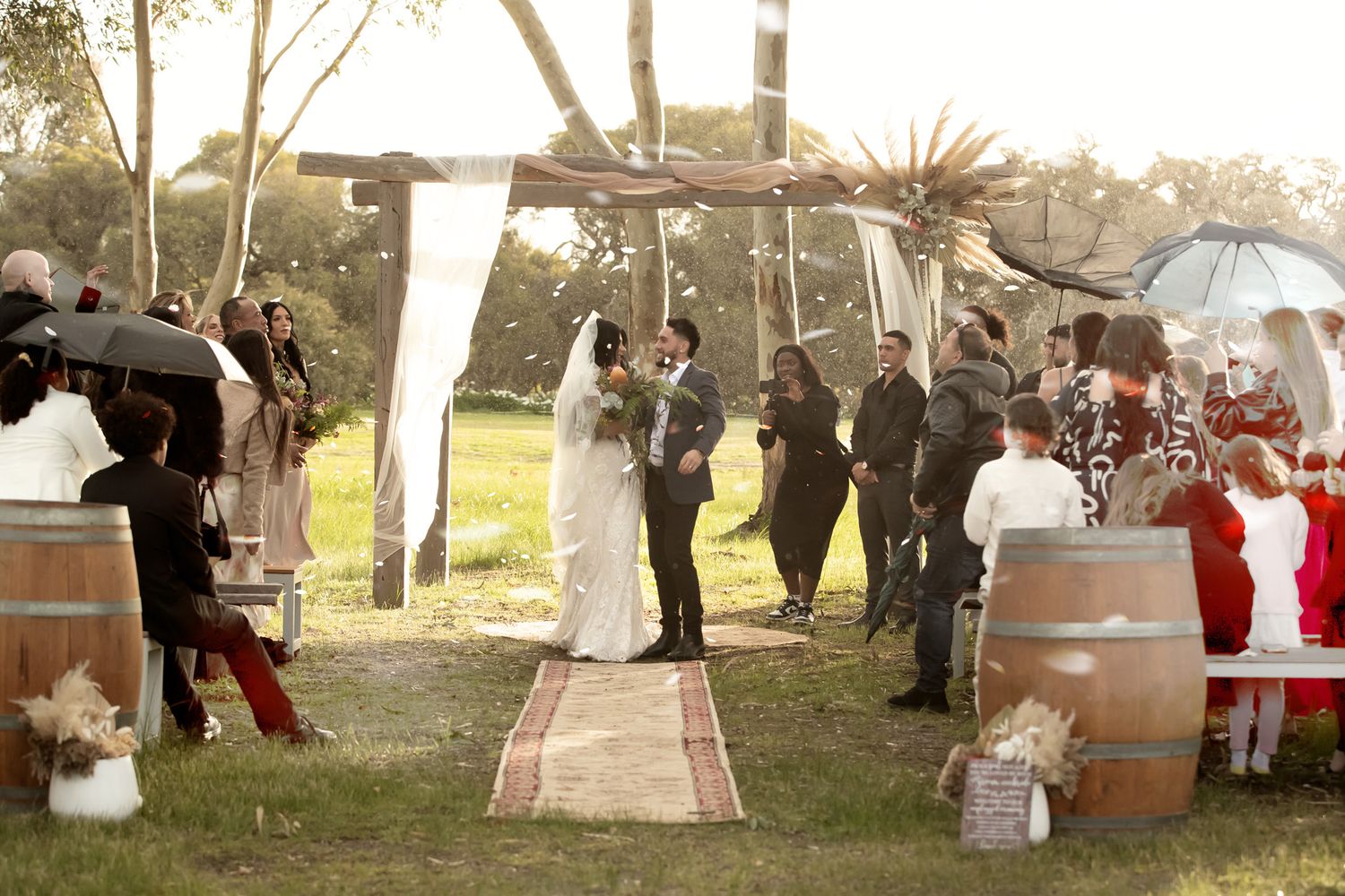 Outdoor rustic wedding ceremony with couple at altar under wooden arch, wine barrel decorations, and guests on green lawn.