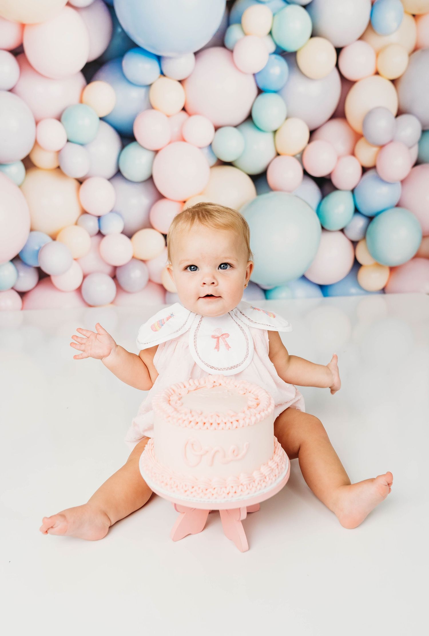 Baby sitting behind a white cake on a gold cake stand during a first birthday cake smash photo session.