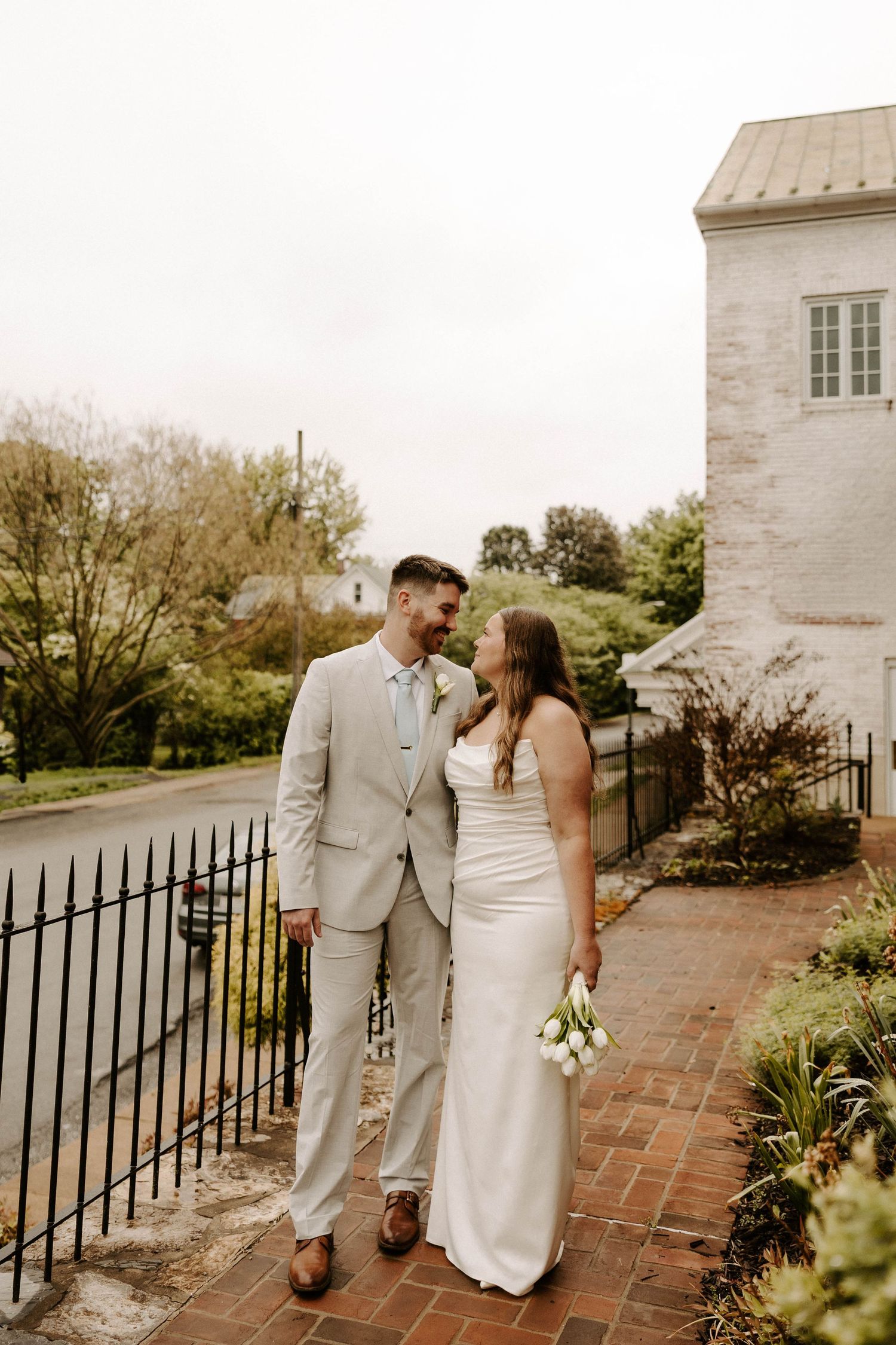 Wedding flat lay with RSVP card, couple photo, lace ribbon, floral bouquet, and gold jewelry on green grass.