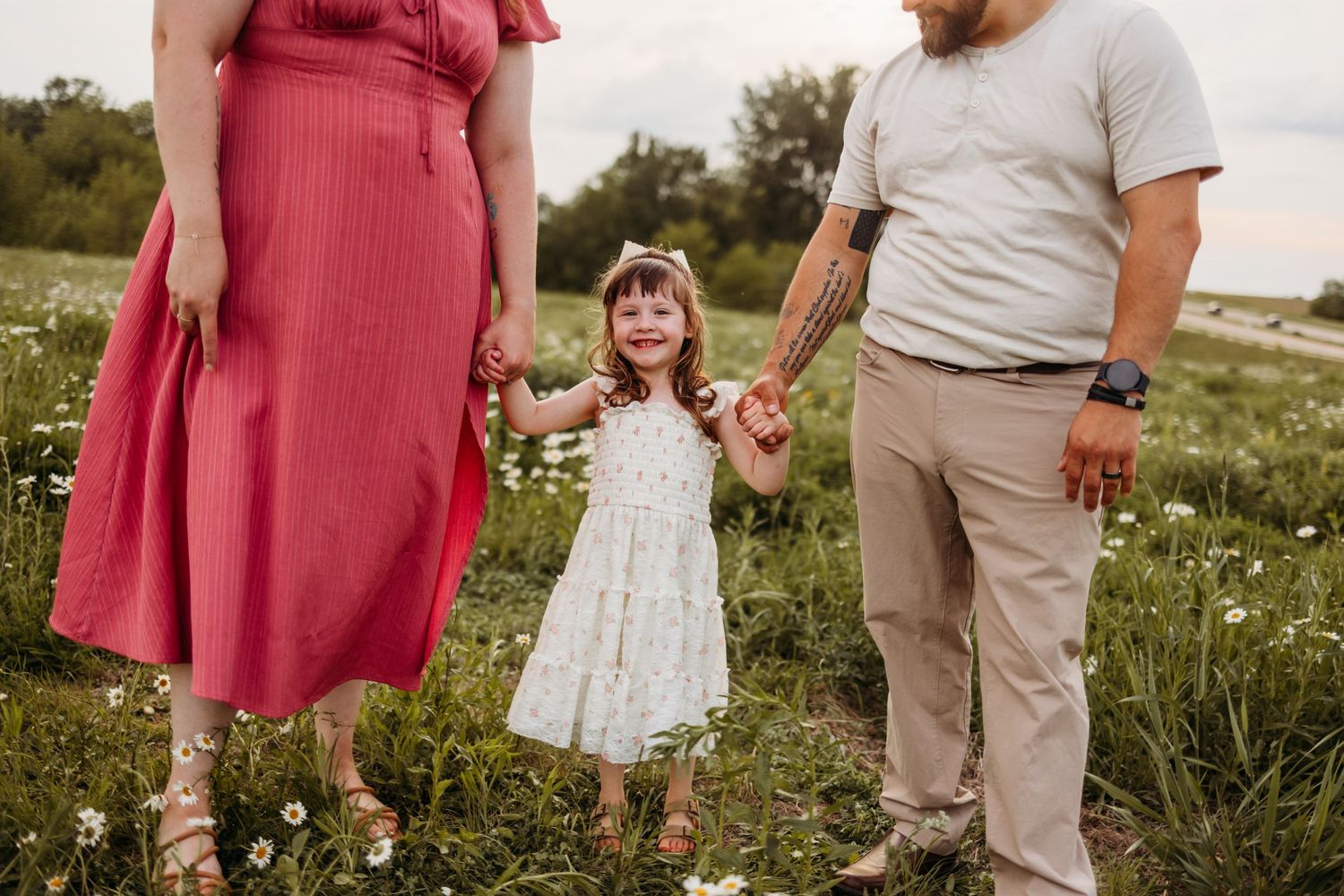 A young family holds hands in a wildflower field, with a smiling little girl in a white dress between her parents.