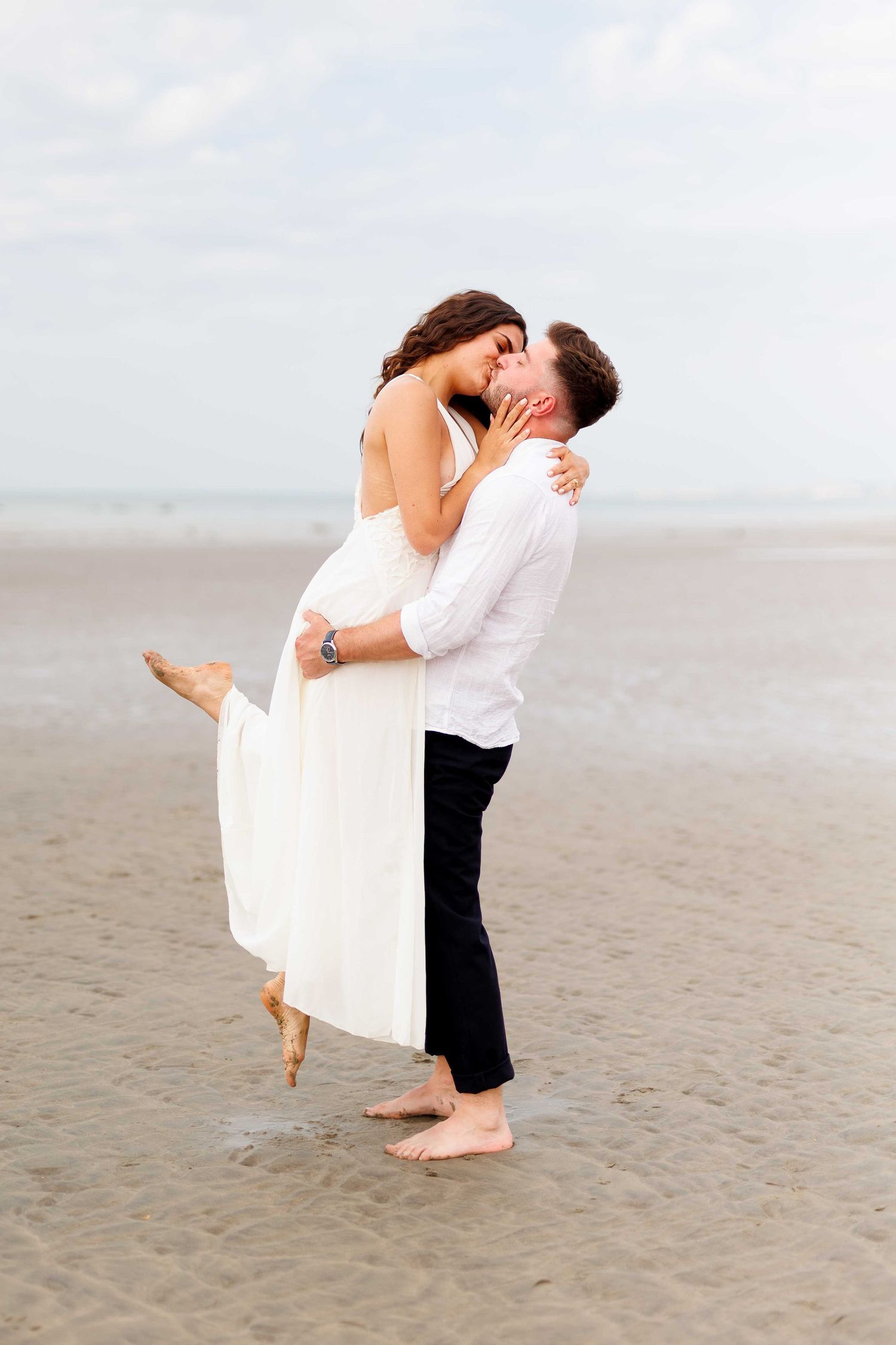 Couple sharing a romantic kiss on the beach, man lifting woman in white dress, barefoot on sandy shore.