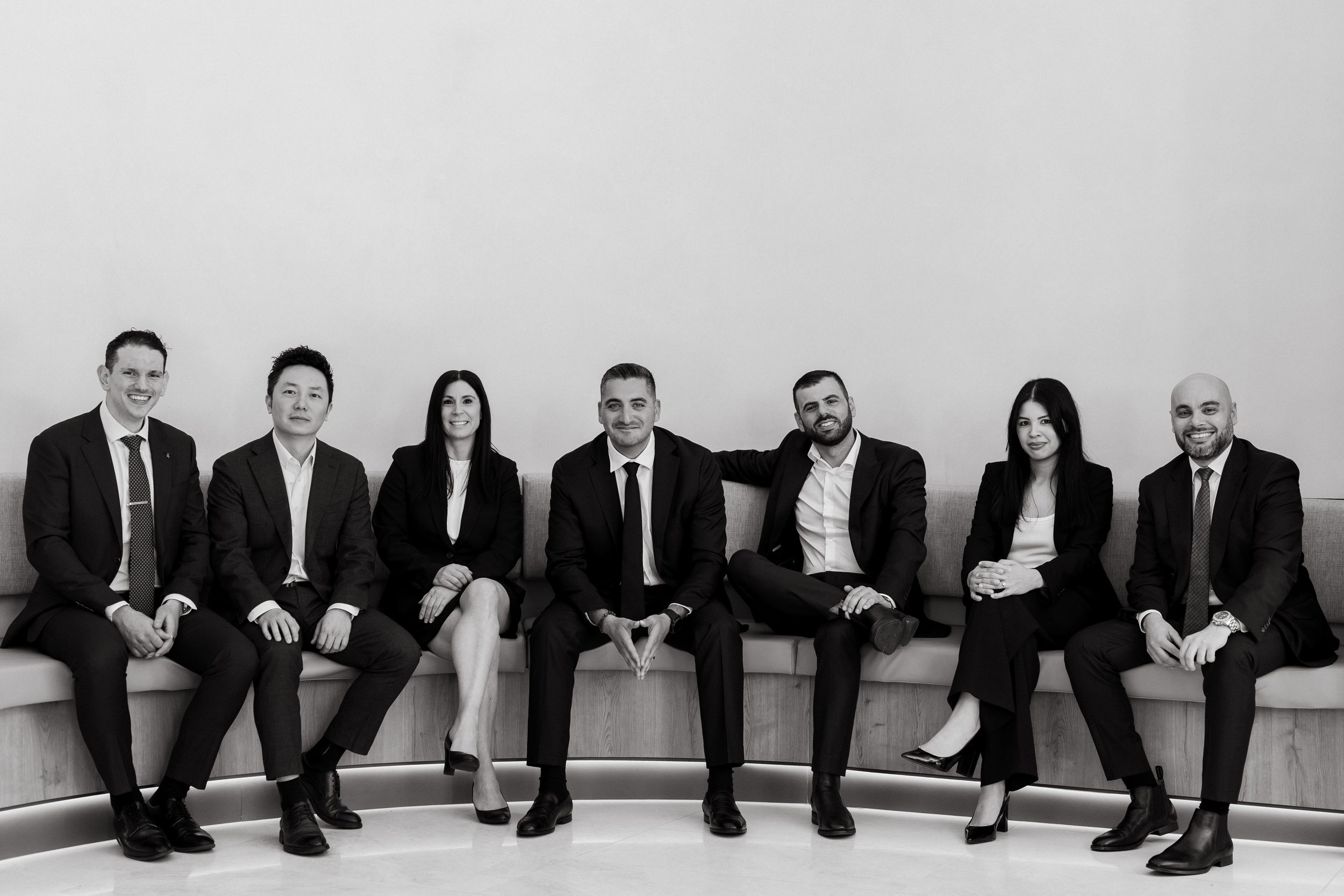 Black and white professional group photo of seven business professionals in formal attire seated on a curved bench.
