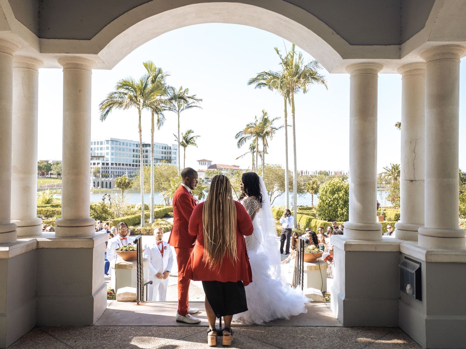 Bride in white gown with lace veil and groom in orange suit pose in lush garden setting on their wedding day.