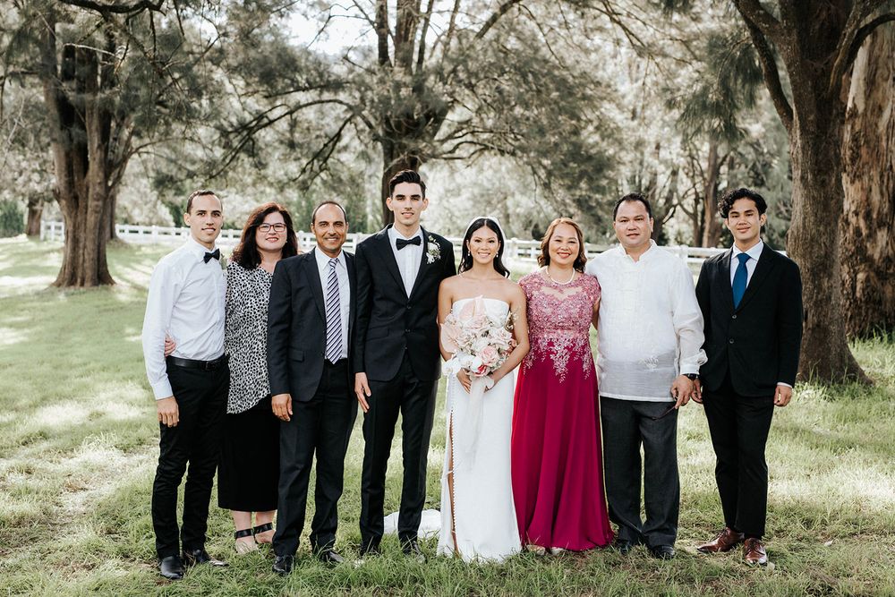 Wedding party group photo with bride, groom, and family posing outdoors under large oak trees.
