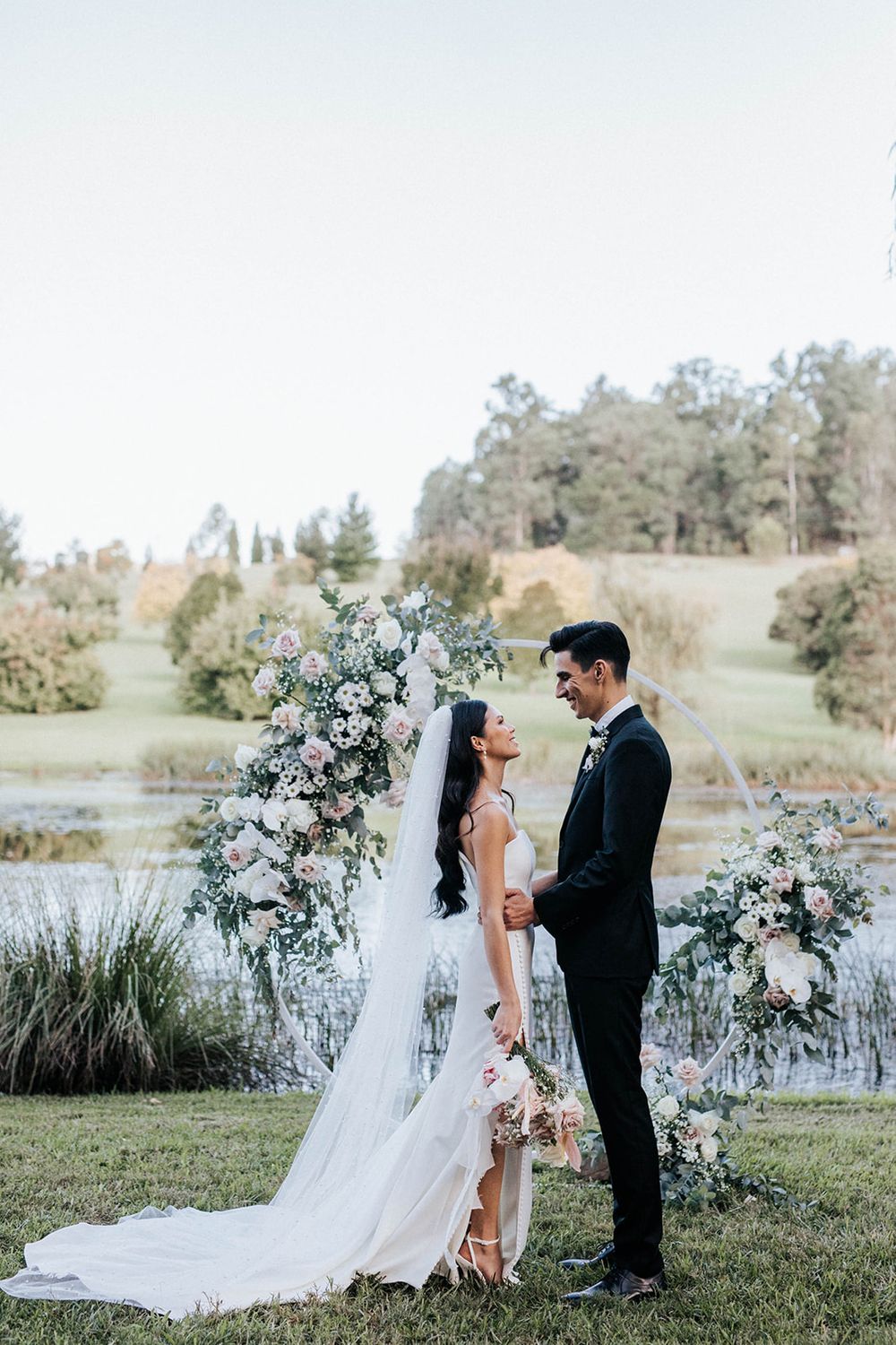 Bride and groom share a romantic moment at an outdoor wedding ceremony with floral arch and scenic lake backdrop.