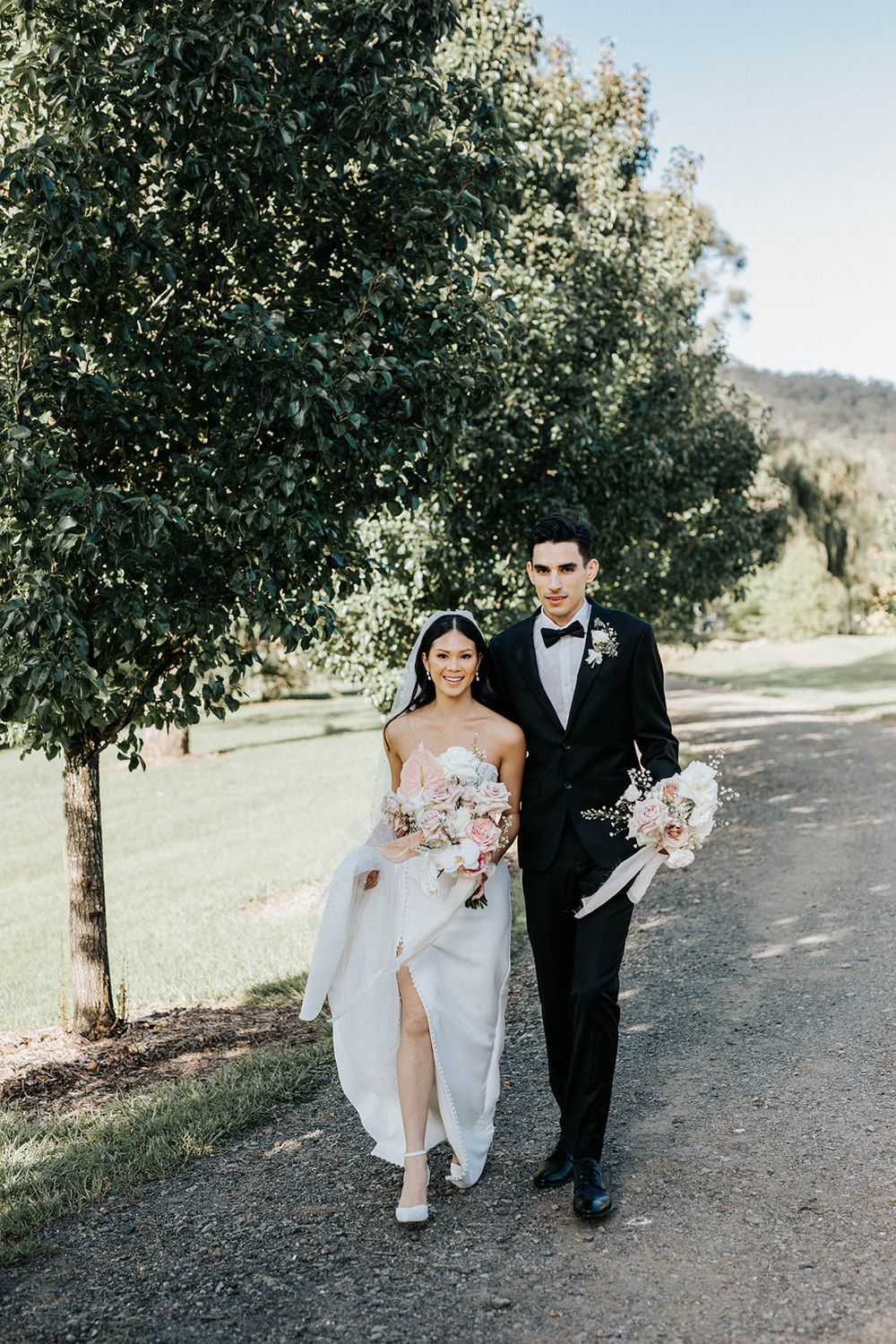 Bride and groom walking hand in hand along a tree-lined path, bride holding pink bouquet in white gown.