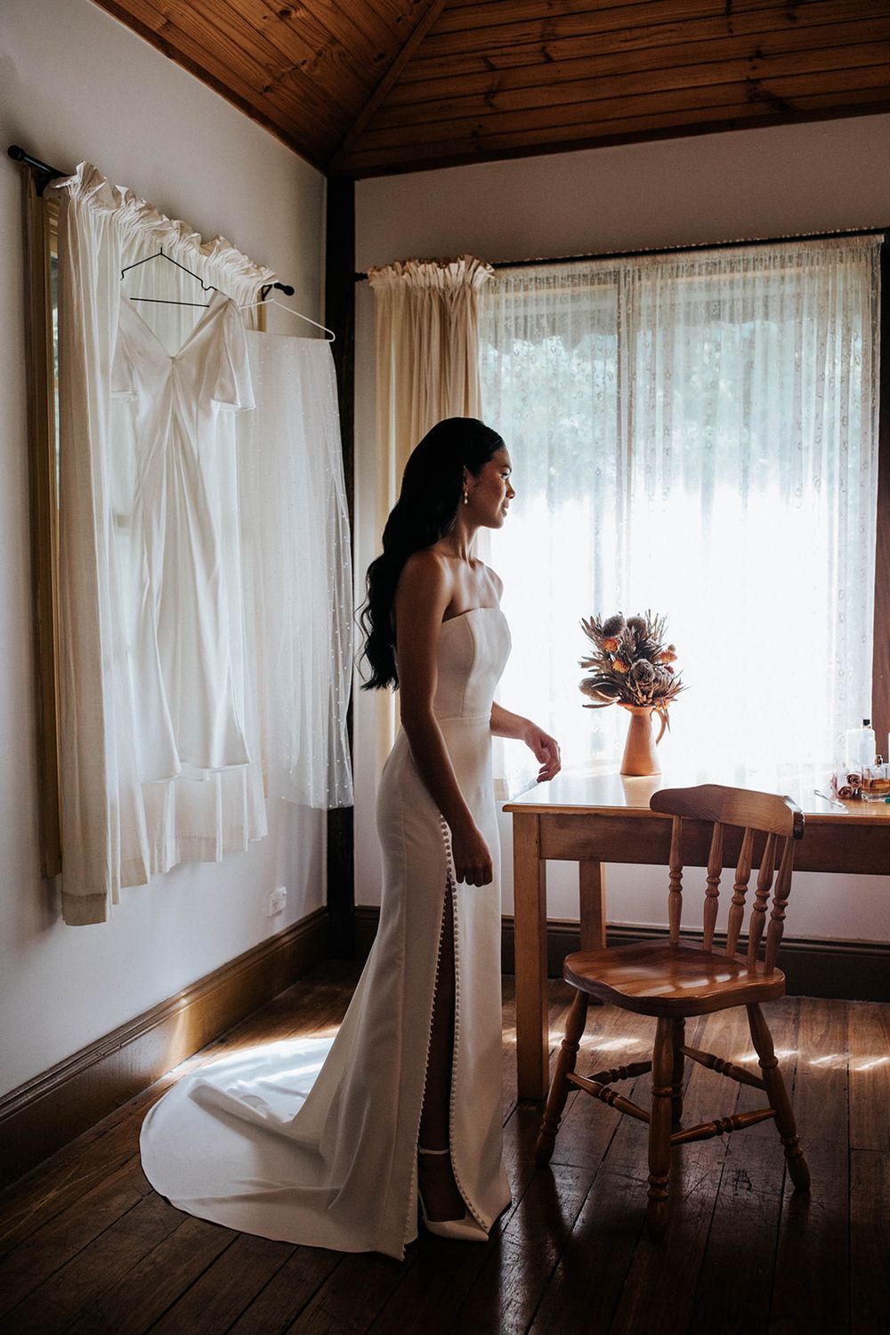 Bride in elegant white mermaid gown stands by sunlit window in rustic cabin room with dresses hanging on wall.