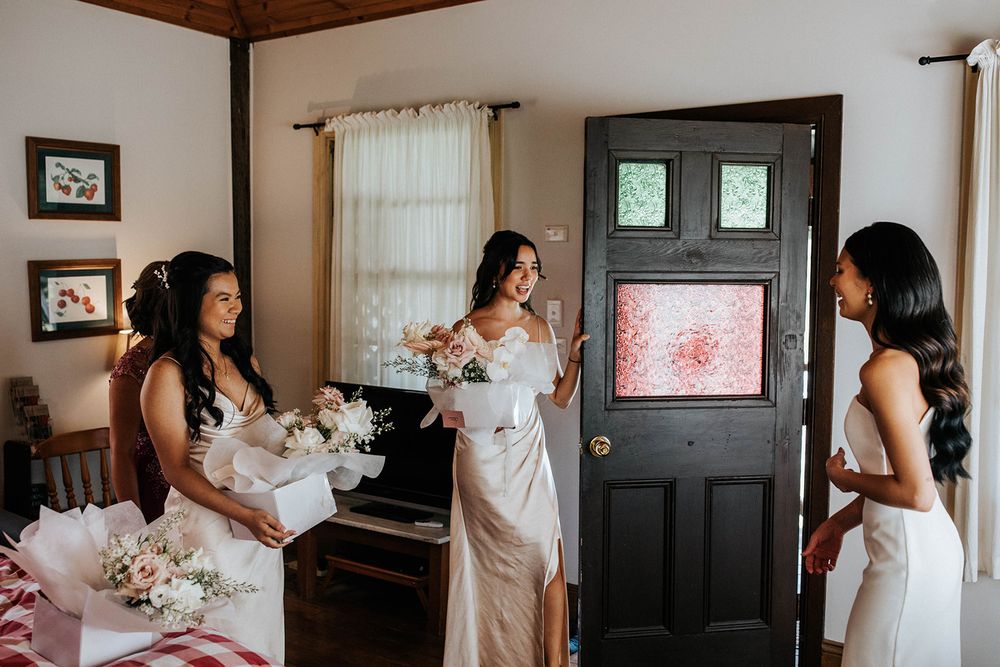 Three bridesmaids in white dresses exchange bouquets of flowers in a rustic room before a wedding ceremony.