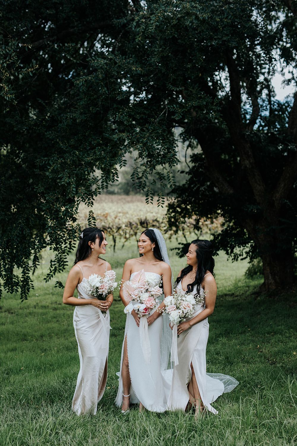 Bride in white gown with veil stands between two bridesmaids in ivory dresses holding bouquets outdoors.