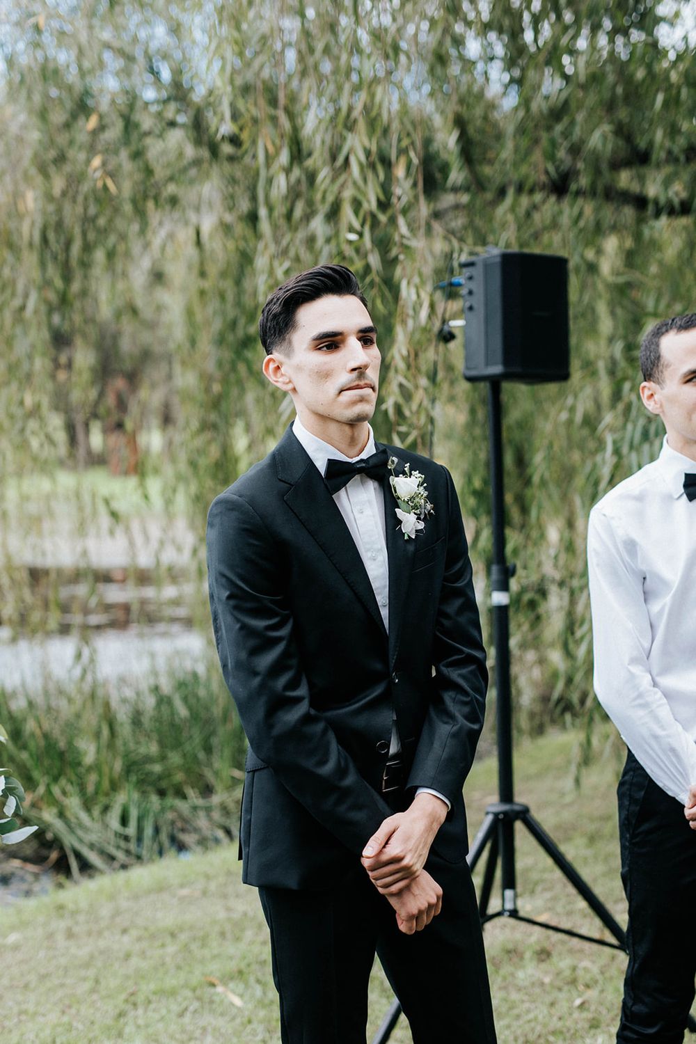 Groom in black tuxedo with boutonniere stands at outdoor wedding altar near willow tree, clasping hands.