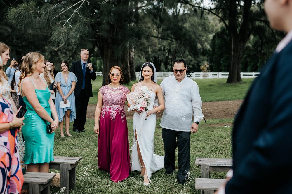 Bride walks down outdoor aisle flanked by parents, guests seated on wooden benches, lush green trees in background.