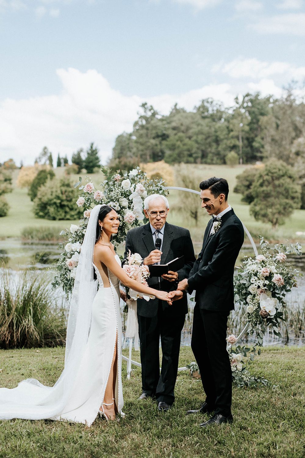 Bride and groom exchange vows outdoors with officiant, floral arch, and scenic pond backdrop on their wedding day.