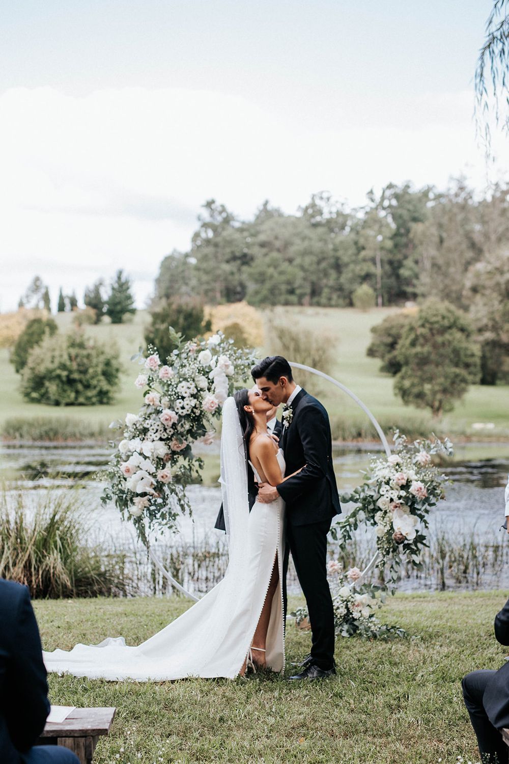 Bride and groom share a kiss at outdoor wedding ceremony with floral arch and scenic lake backdrop.
