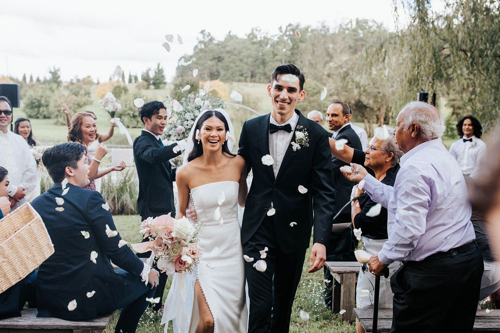 Happy newlyweds walk down the aisle as guests shower them with white flower petals at outdoor wedding ceremony.