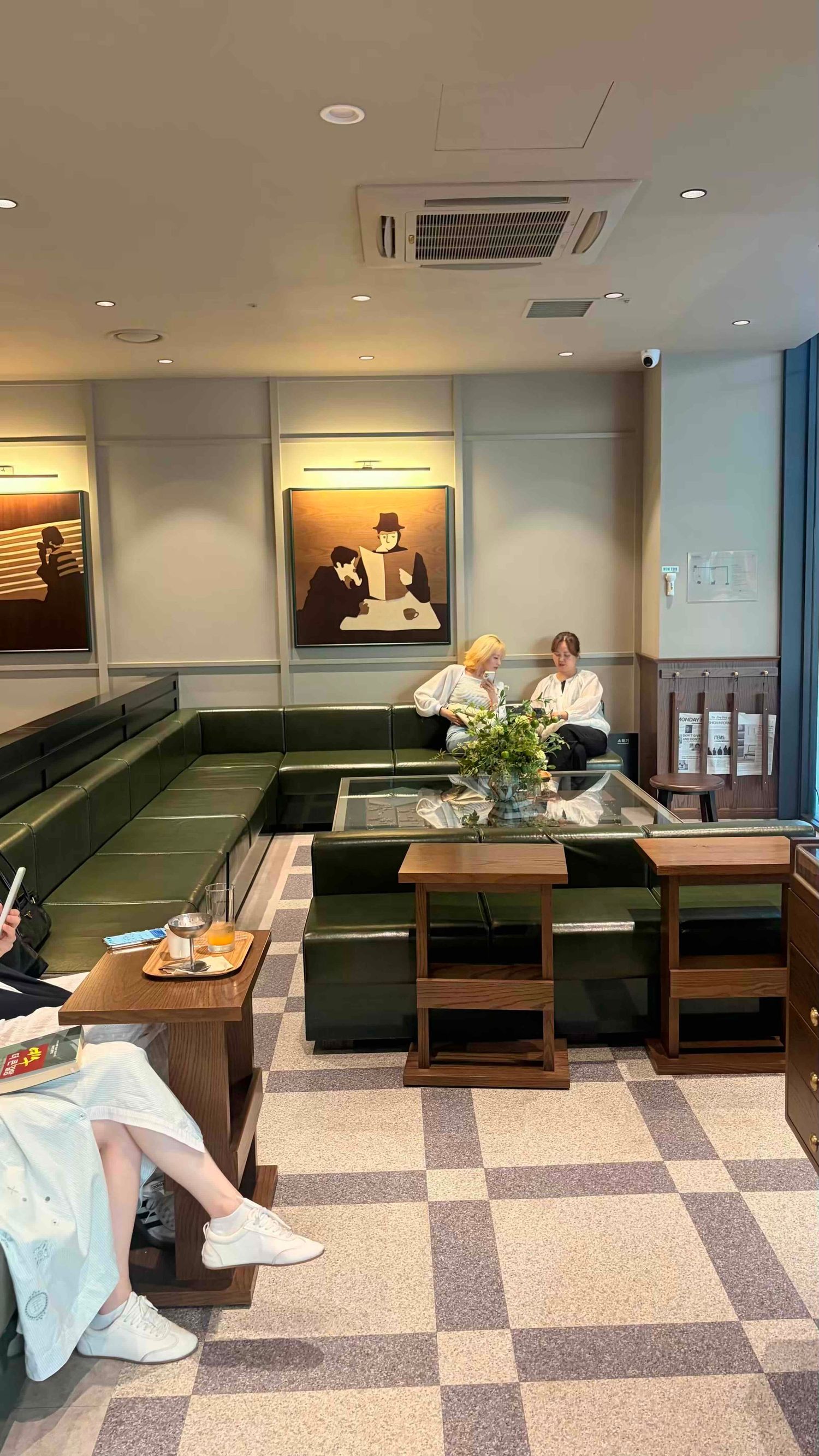 Modern cafe interior with green leather booths, checkered floor, artwork on walls, and patrons relaxing at wooden tables.