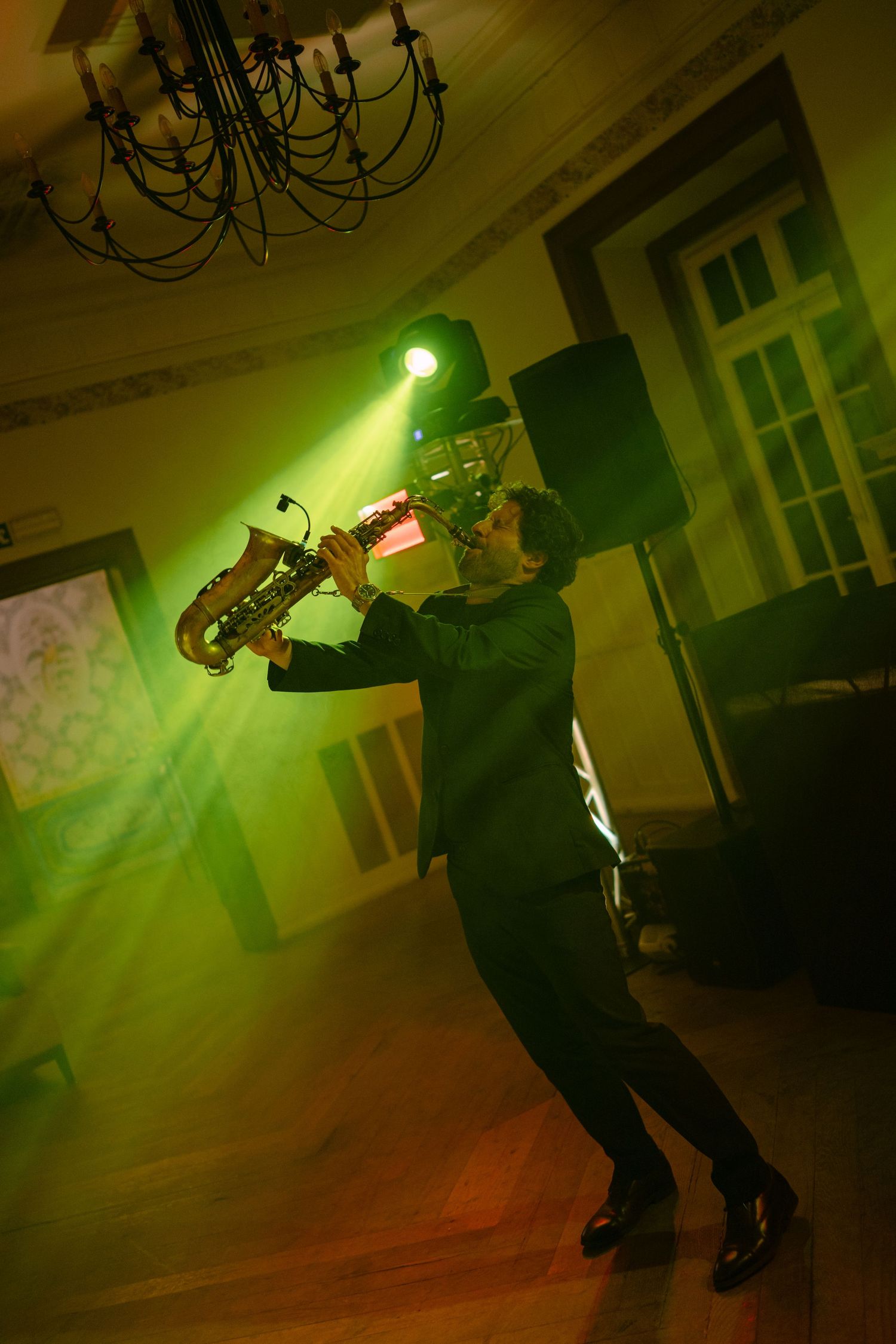 Saxophonist performing under green stage lights in elegant venue with chandelier overhead.