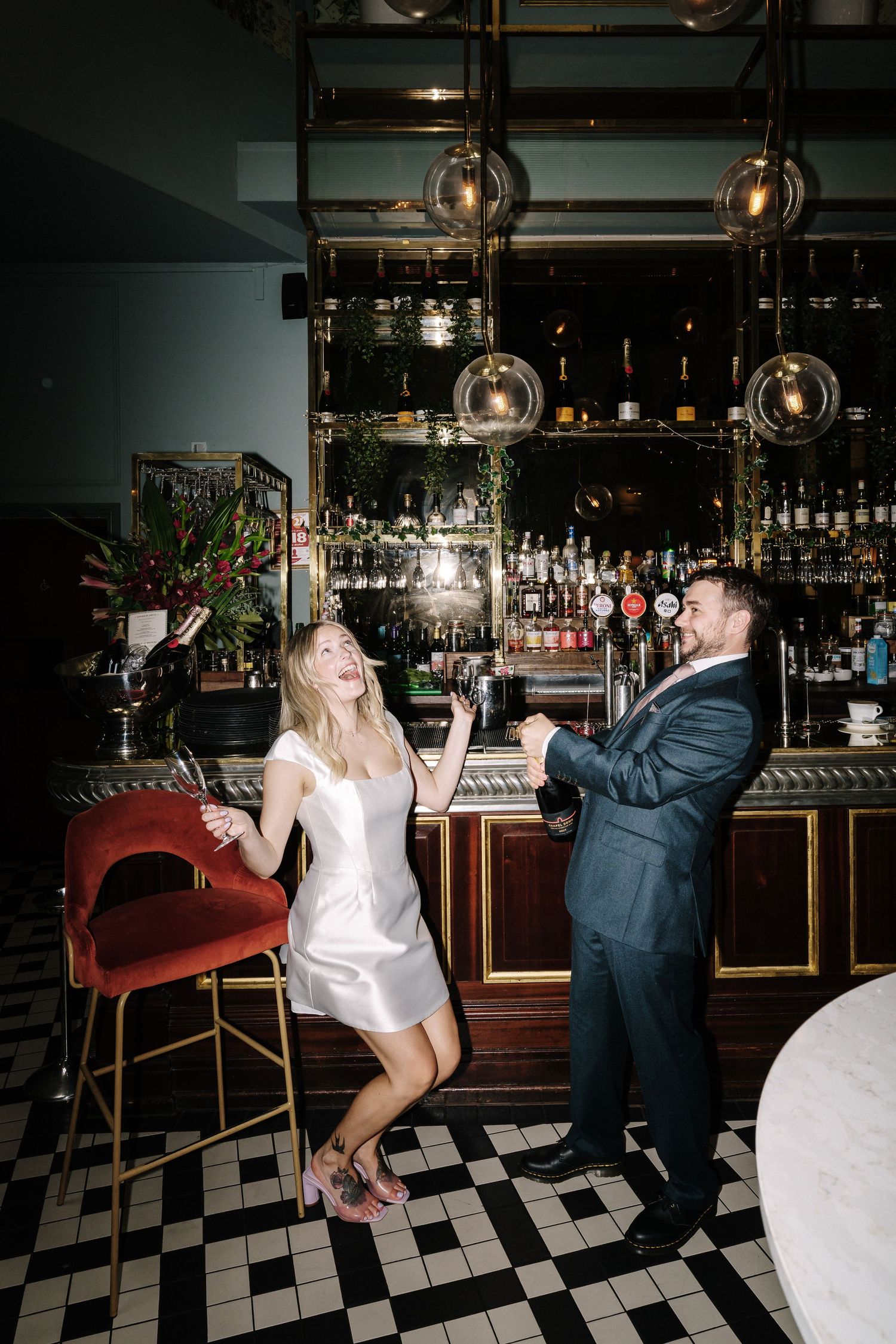 A laughing couple dances joyfully at an elegant bar with checkered floors, globe lights, and a well-stocked backdrop.