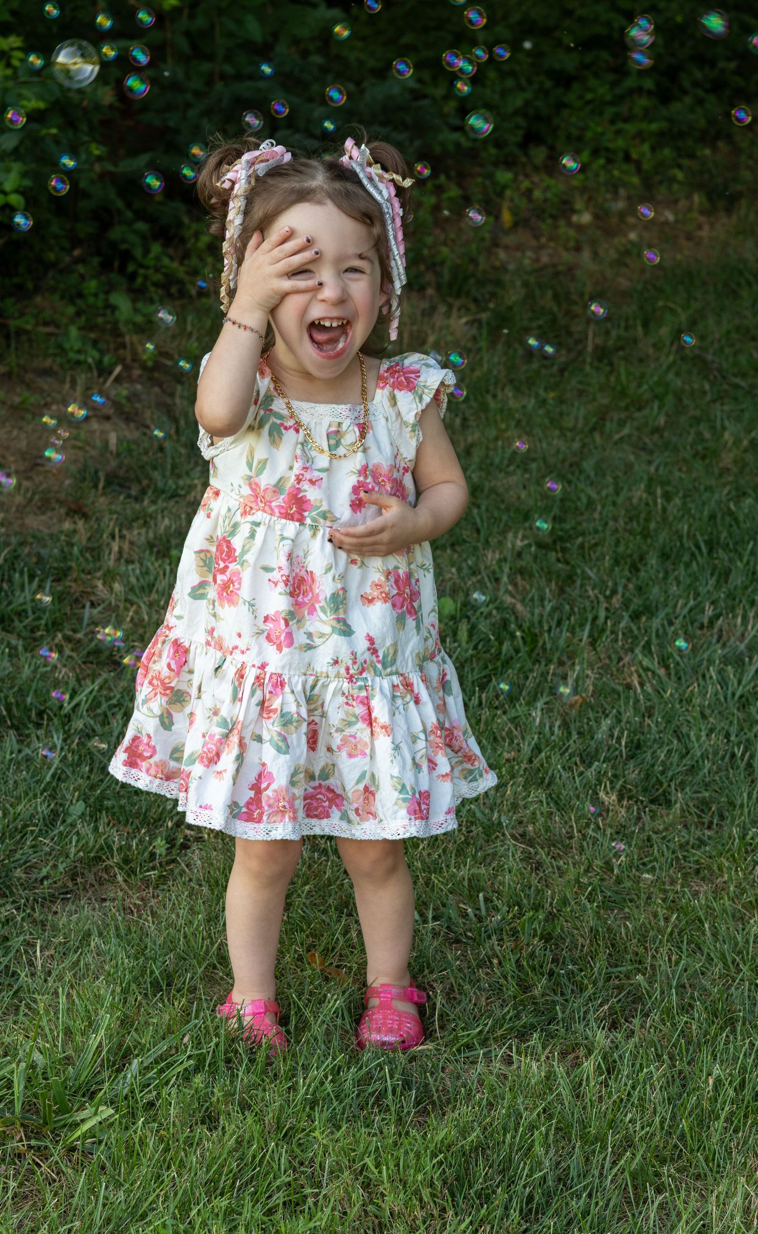 A joyful toddler girl in a floral dress laughs while playing with bubbles outdoors on green grass.