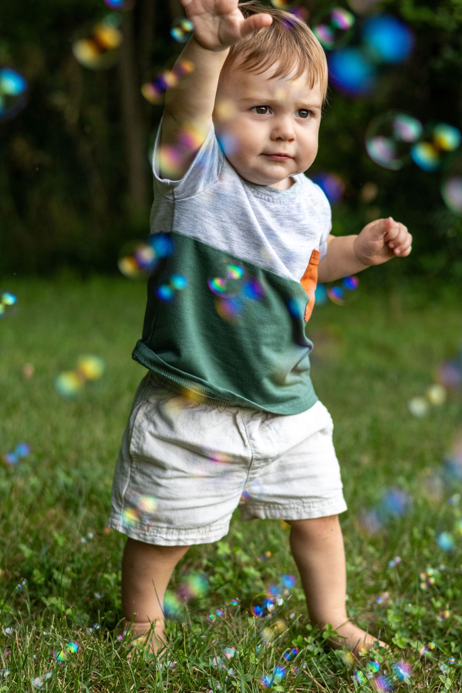 Toddler boy in colorful shirt and white shorts reaching for soap bubbles on green grass outdoors.