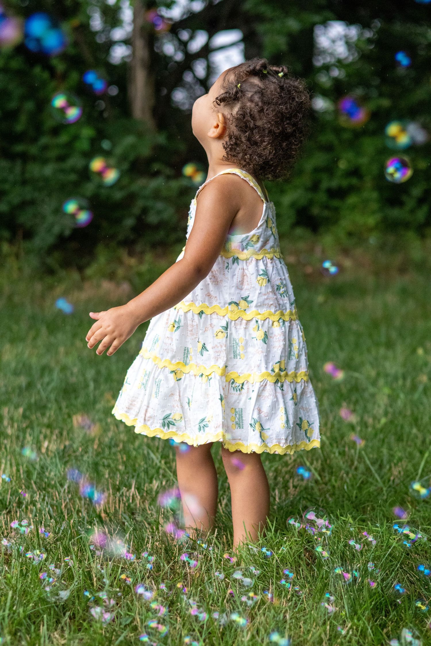 Young girl in white floral dress playing with colorful soap bubbles on green grass outdoors.
