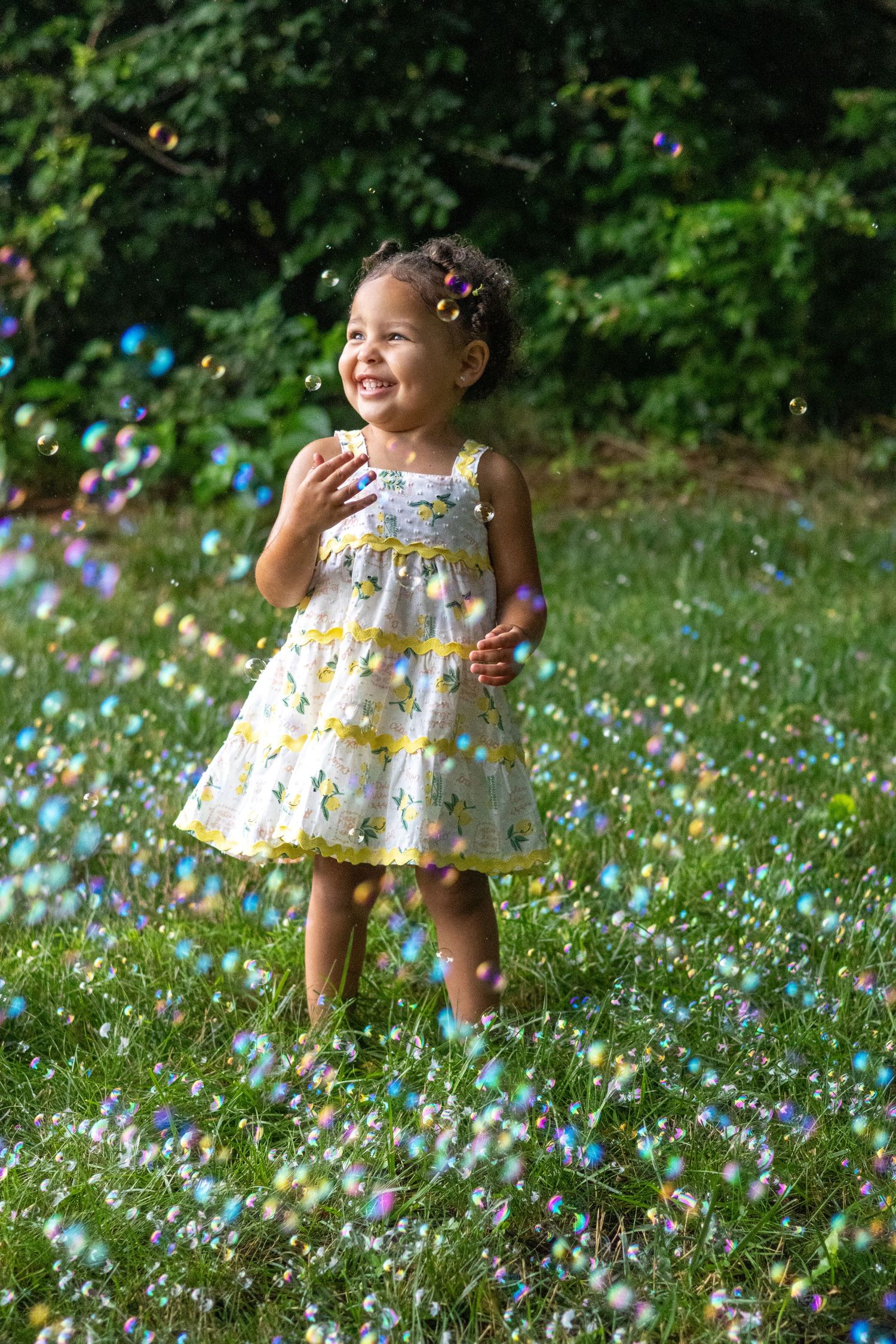 A joyful toddler girl in a floral dress laughs while playing with colorful bubbles on a sunny green lawn.