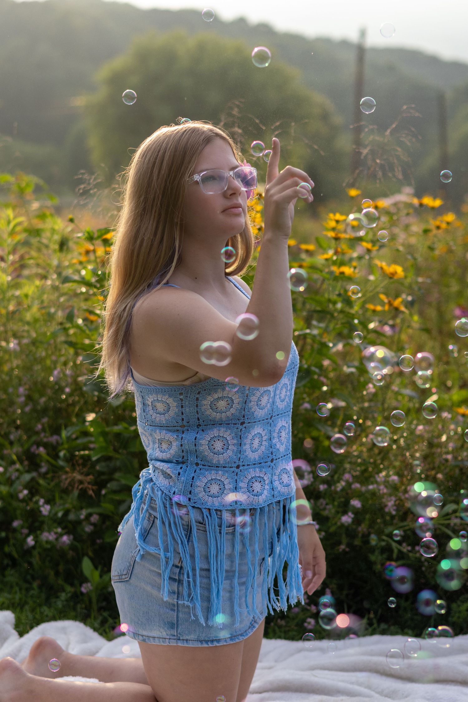 Young woman in crochet top and denim shorts playing with soap bubbles in a sunny garden with wildflowers.