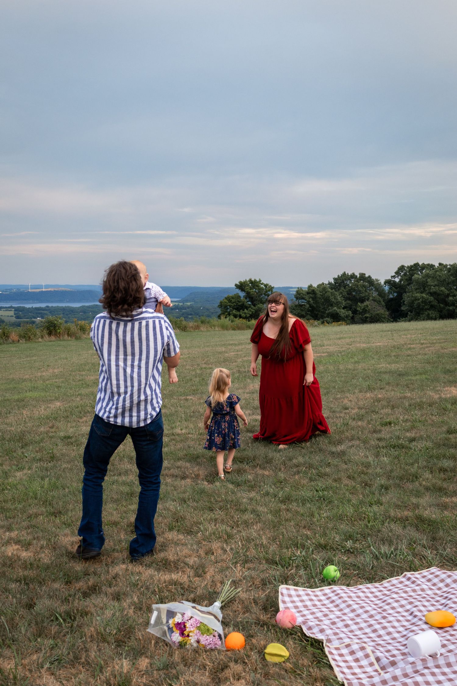Family enjoying a outdoor picnic on a grassy hill, with a man holding a baby, a young girl, and a woman in a red dress.