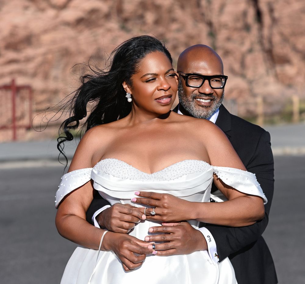 A joyful couple in wedding attire embraces outdoors, bride in white gown, groom in suit, red rocks background.