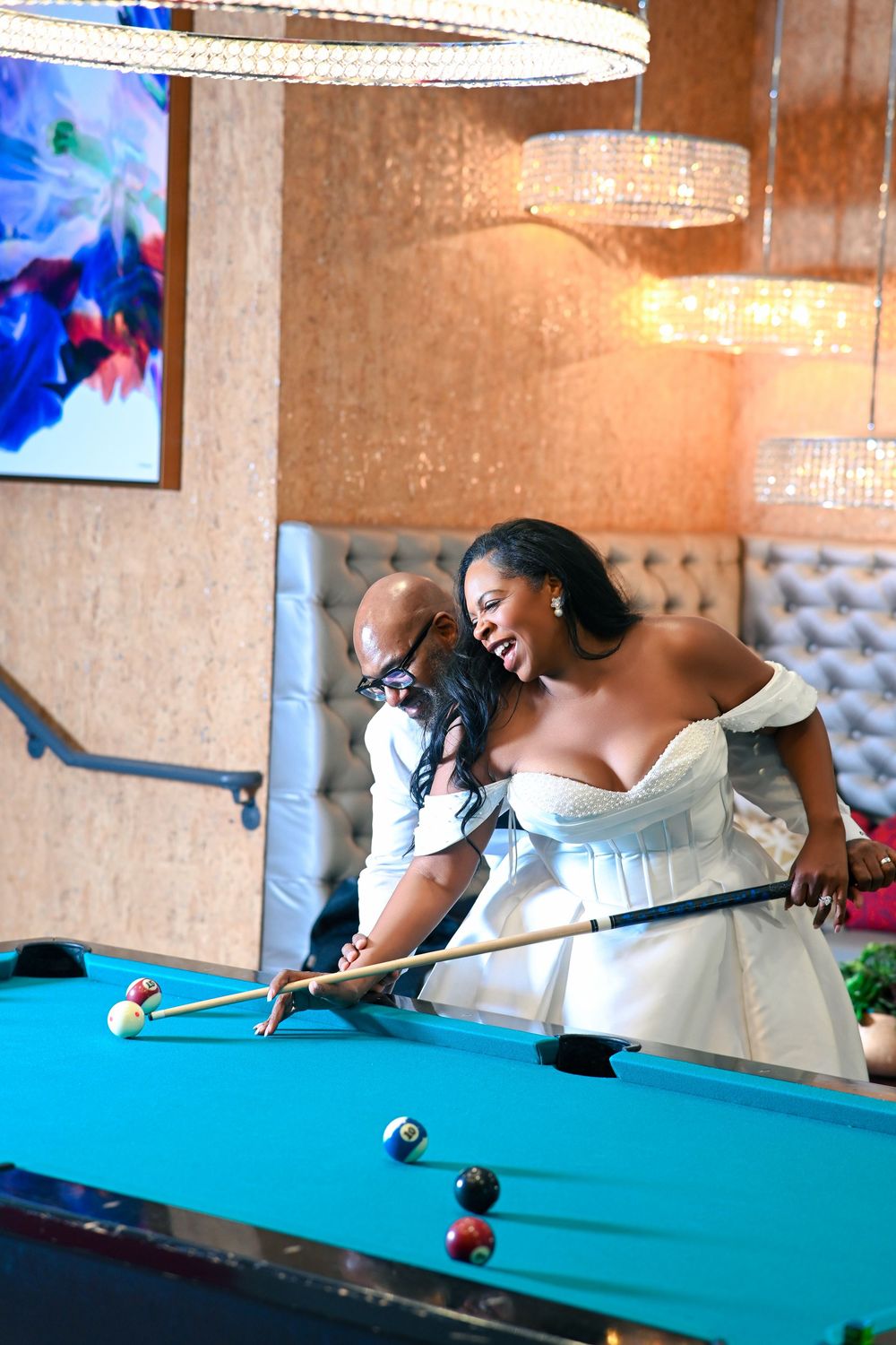 Bride in white off-shoulder gown playing pool with groom at elegant venue with crystal chandeliers.