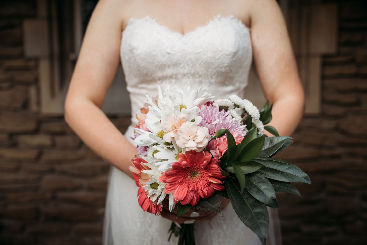 Bride in white lace strapless gown holding colorful wedding bouquet with pink gerbera daisies and white flowers.