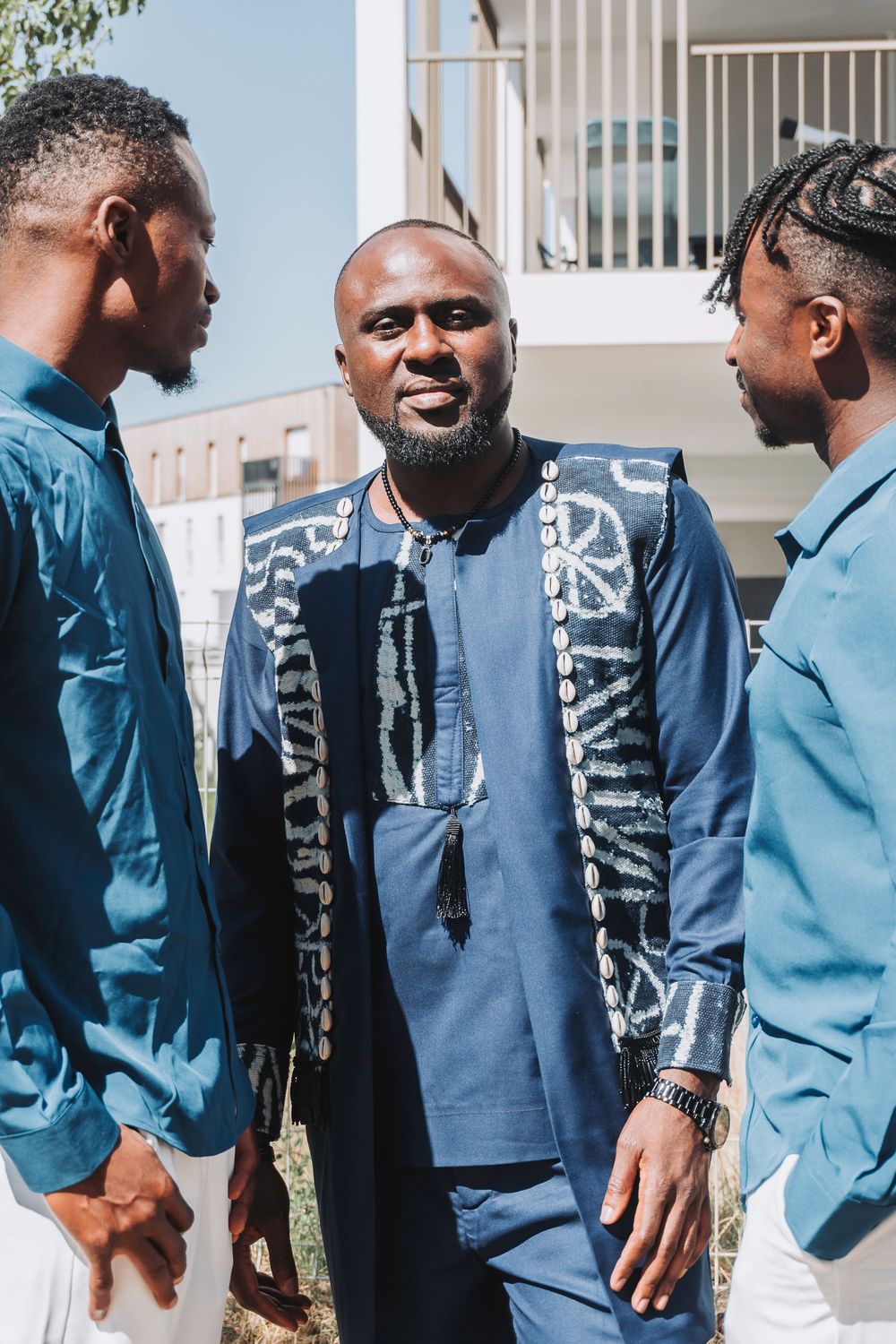 Three Black men in blue outfits, center man wearing traditional African attire, conversing outdoors on a sunny day.