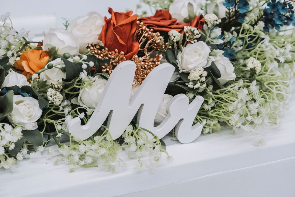 White Mr. sign surrounded by wedding floral arrangement with red, white, and orange roses and greenery.
