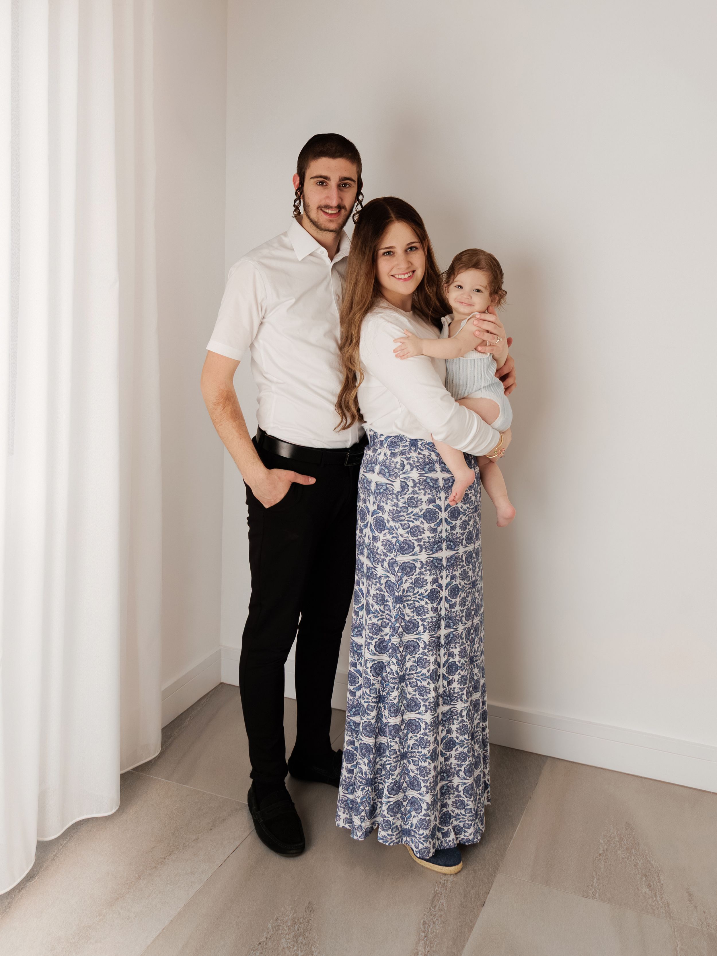Young family of three posing together, man in white shirt, woman in floral maxi skirt holding a toddler.
