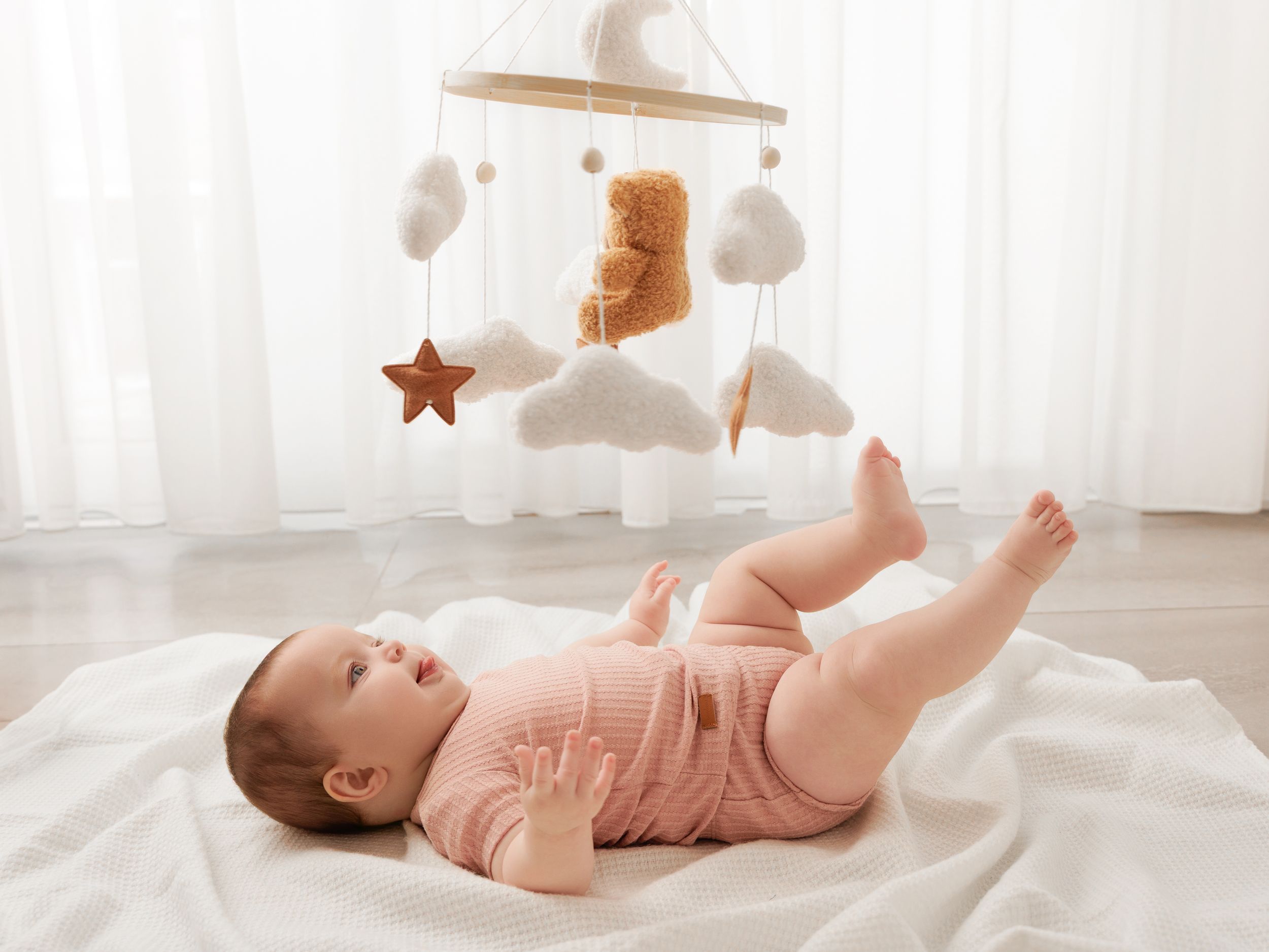 Baby lying on white blanket gazing at a nursery mobile with teddy bear, stars, and cloud decorations.