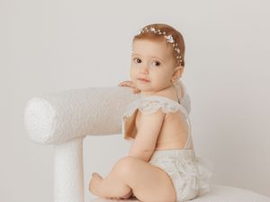 Adorable baby girl in white lace romper and floral headband poses on white chair for professional portrait.