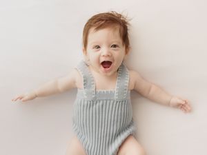 Baby wearing light blue striped romper lies on white blanket with joyful expression and outstretched arms.